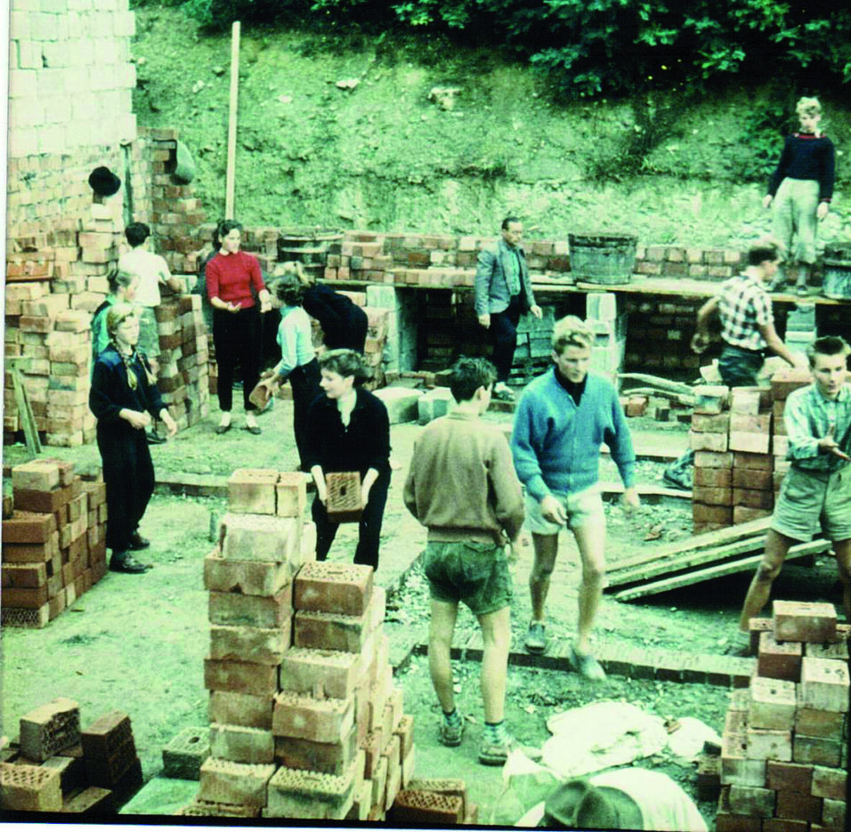 Bauarbeitende Menschen im Ziegelsteinlager, umgeben von gestapelten Ziegeln und Halbfertigen Wänden.