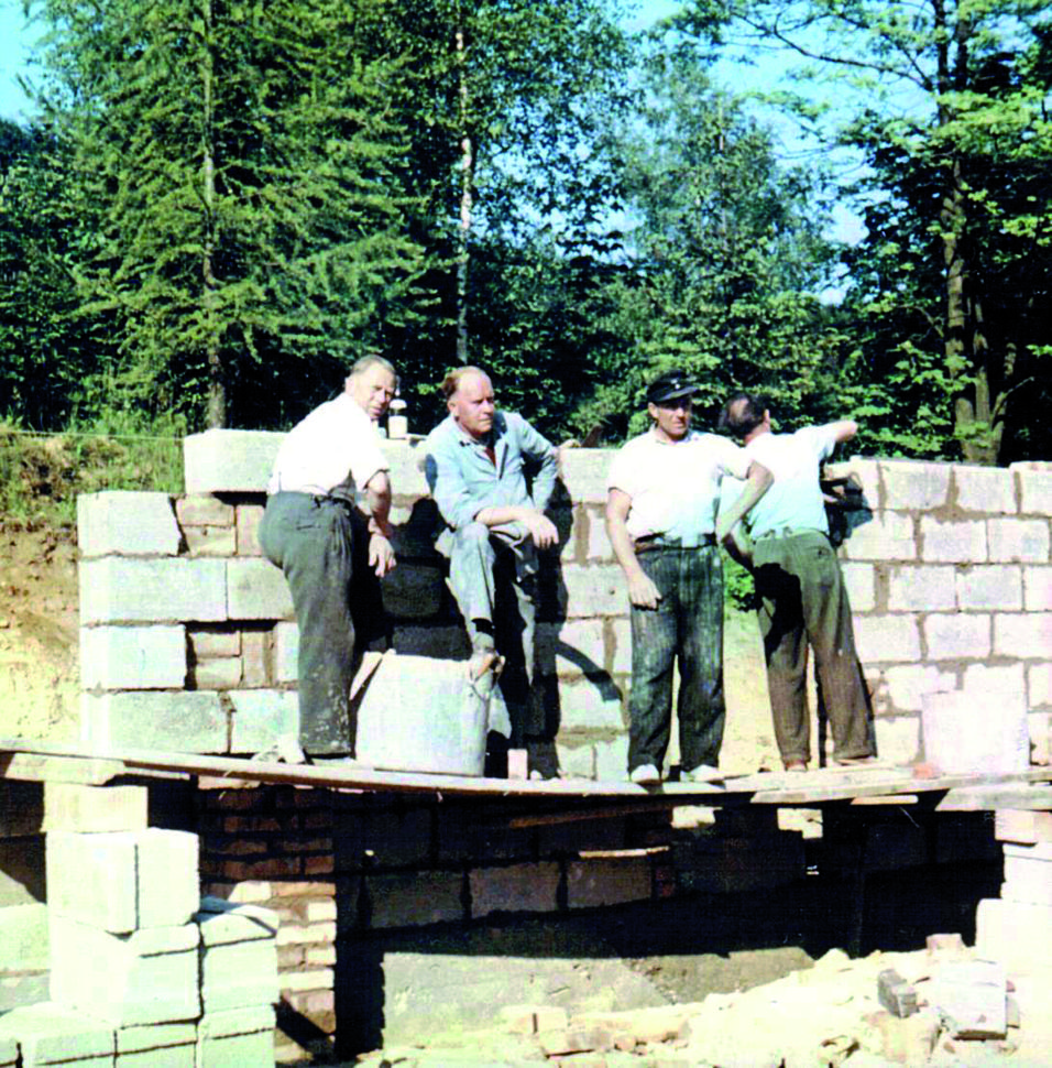 Fünf Bauarbeiter stehen auf einer Rohbaustelle, umgeben von Steinen und Bäumen, während sie an einer Mauer arbeiten.