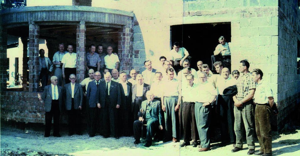 Gruppe von Männern unterschiedlicher Altersgruppen vor einem Rohbau, einige stehen, andere auf einem Balkon.