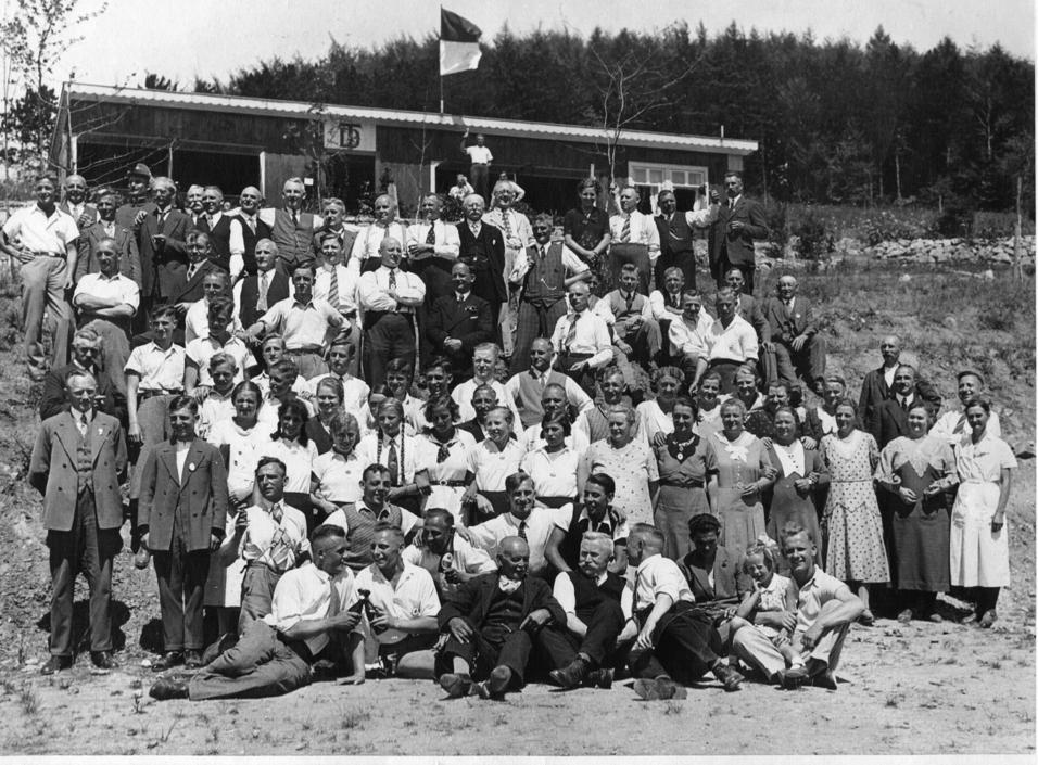Gruppenfoto von über 70 Personen vor einem Gebäude, einige tragen historische Kleidung und eine Fahne weht im Hintergrund.