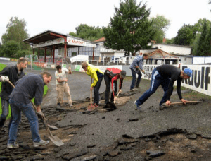 Gruppe von Menschen arbeitet gemeinsam daran, den Untergrund eines Sportplatzes mit Schaufeln zu bearbeiten.