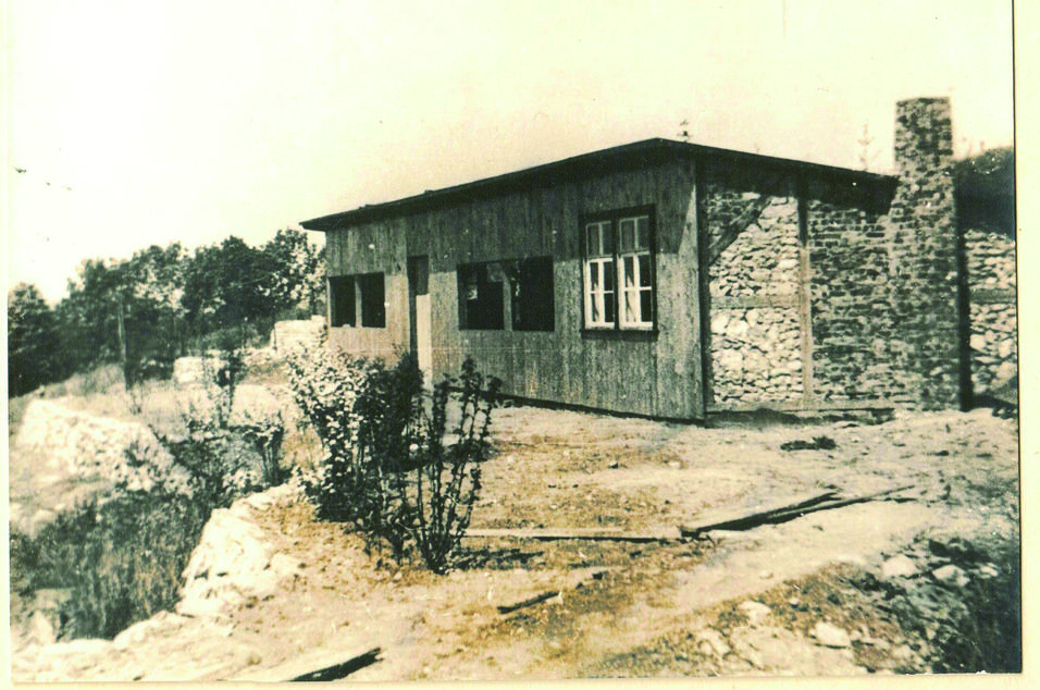 Ein einfaches, einstöckiges Gebäude mit Holzfassade und Steinmauer, umgeben von Büschen und Bäumen.