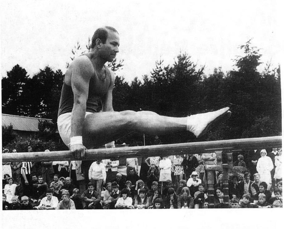 Turner Dieter Herzog in der Übung am Langbarren, während einer Wettkampfveranstaltung mit Zuschauern im Hintergrund.