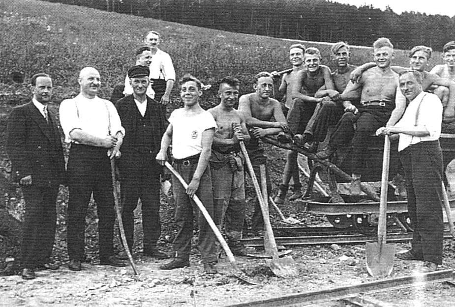 Gruppenfoto von Arbeitern mit Schaufeln, einige tragen Arbeitskleidung, auf einer Baustelle im Freien.