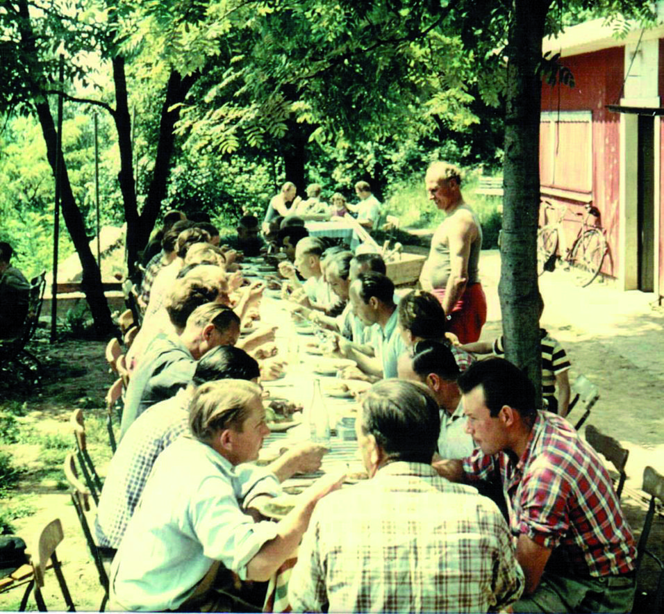 Gruppierung von Männern an einem langen Tisch im Freien, beim Essen unter Bäumen, mit einem roten Gebäude im Hintergrund.