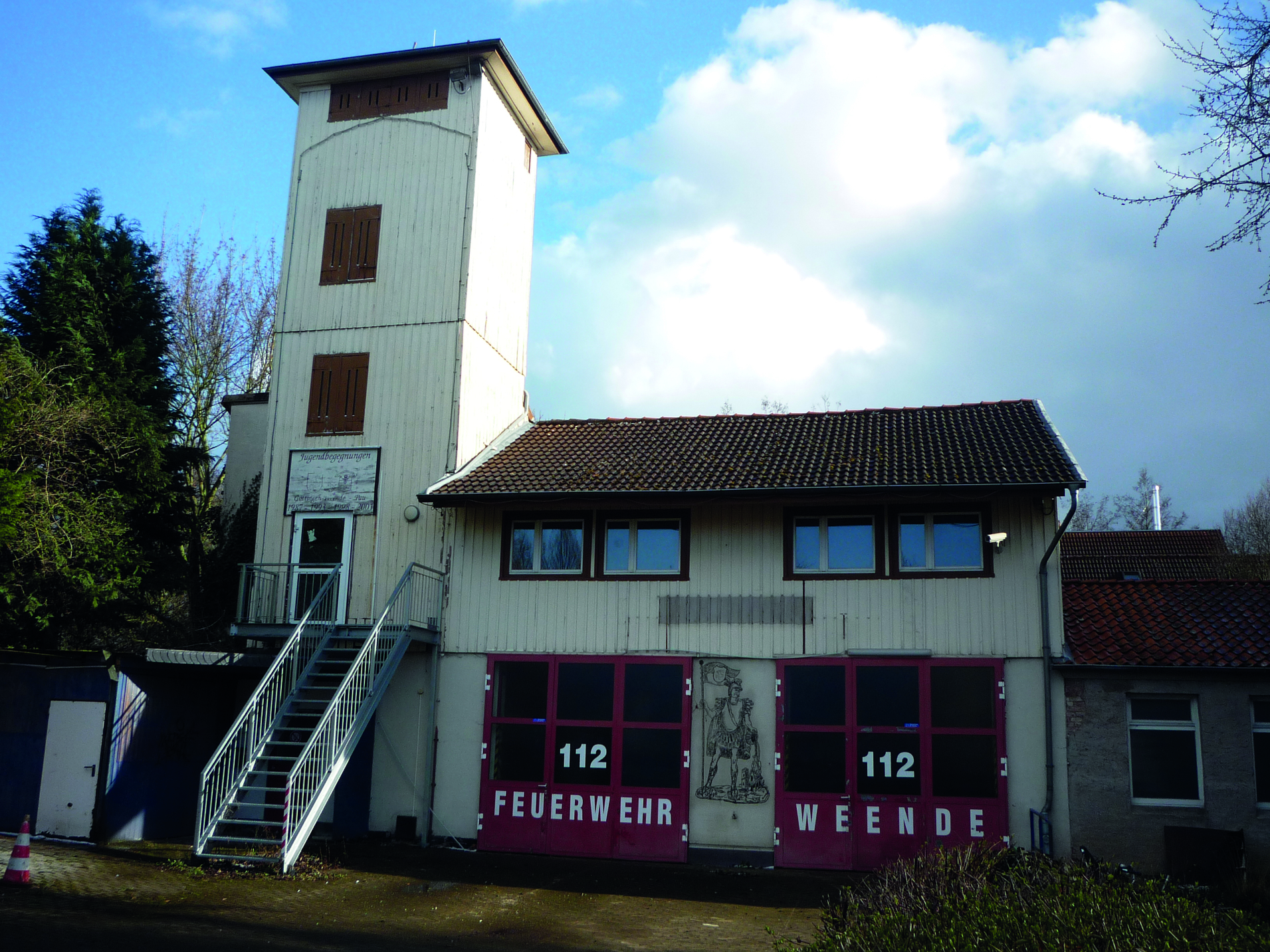 Feuerwehrgeb&auml;ude in Weende mit Aussichtsturm und gro&szlig;en roten Garagentoren. Wolkiger Himmel im Hintergrund.