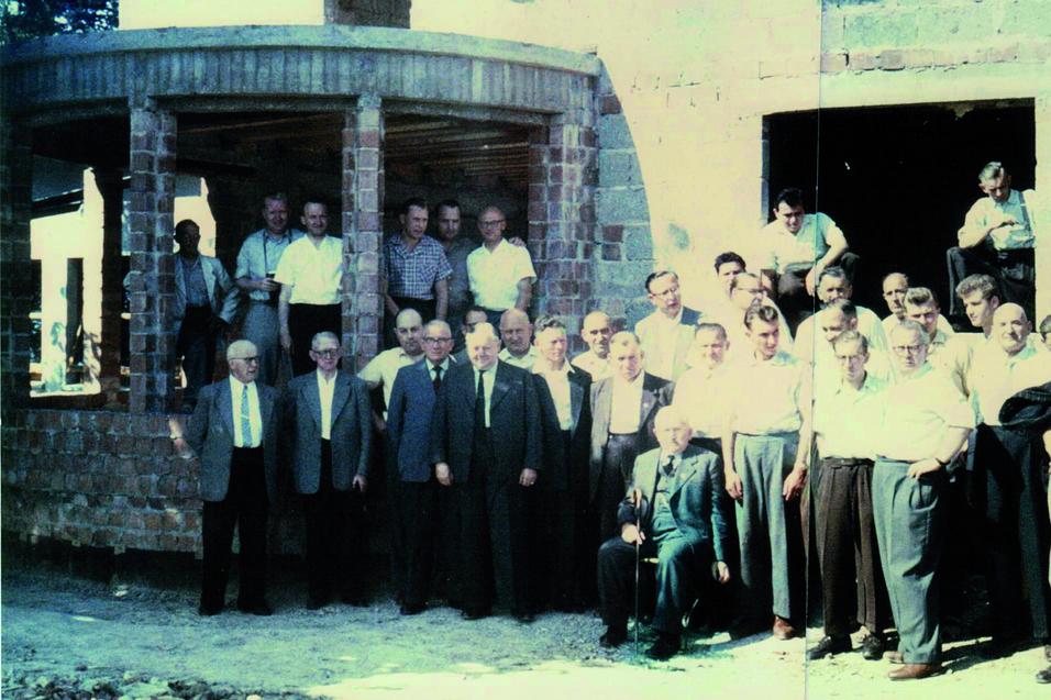Gruppe von M&auml;nnern unterschiedlicher Altersgruppen vor einem Rohbau, einige stehen, andere auf einem Balkon.