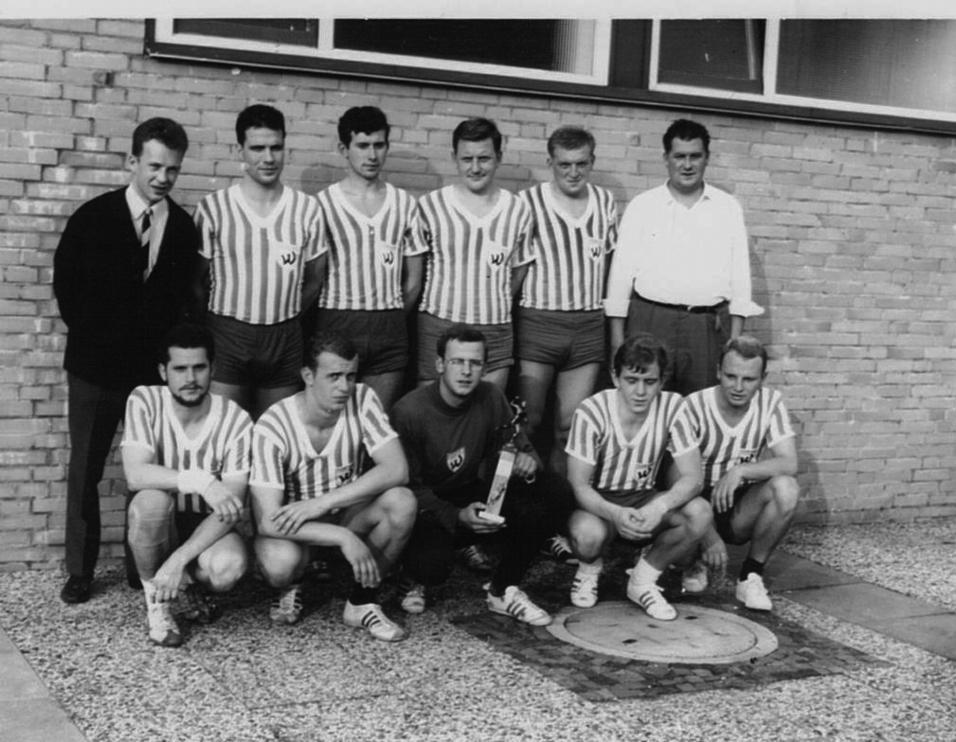 Schwarz-weiß-Foto einer Fußballmannschaft mit Pokal, bestehend aus sieben Spielern und zwei Trainern vor einer Wand.