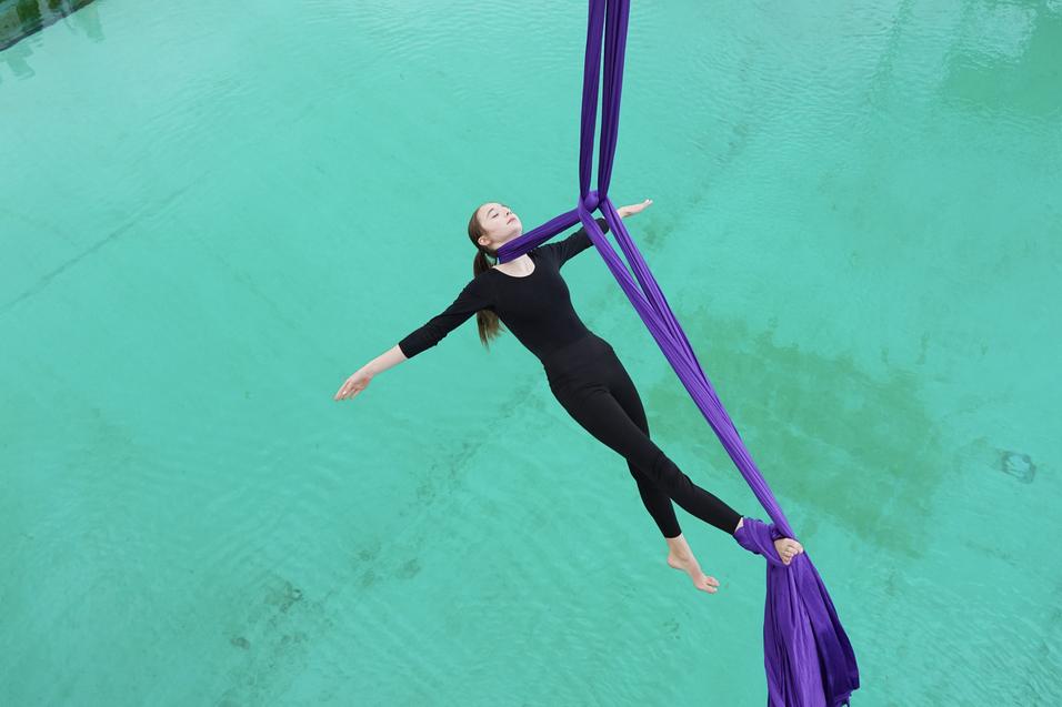 Artistin schwebt an lila T&uuml;chern &uuml;ber einem klaren Wasserbecken, umgeben von einer Zuschauergruppe.
