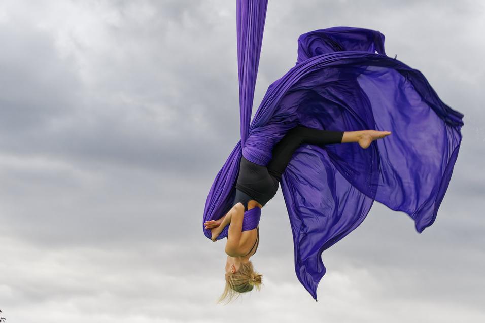 T&auml;nzerin in lila Tuch schwingt kopf&uuml;ber in der Luft vor einem bew&ouml;lkten Himmel.