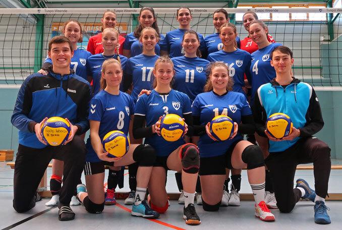 Gruppeneinstellung eines Volleyballteams in blauen Trikots, alle halten einen Volleyball, vor einem Netz in einer Sporthalle.