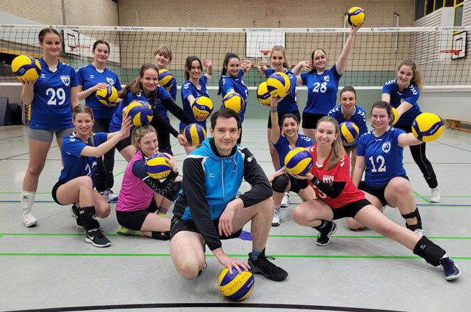 Volleyballmannschaft mit Trainer posiert in Sportkleidung, alle halten Volleyb&auml;lle vor einem Netz in der Halle.