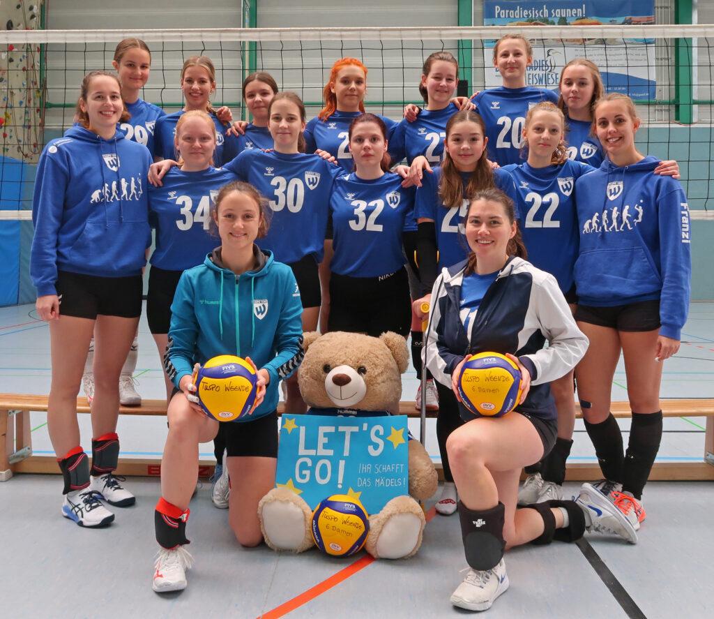 Gruppe von Volleyballspielerinnen in blauen Trikots, posierend mit B&auml;ren und Ball in einer Sporthalle.