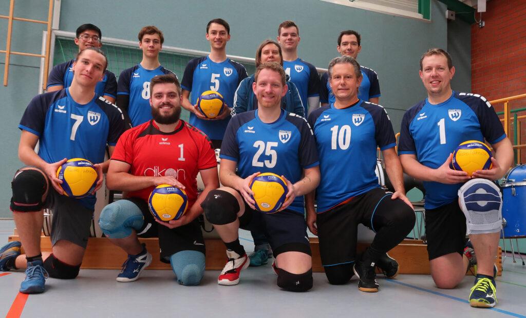 Teamfoto einer Volleyballmannschaft mit bunten B&auml;llen, in blauen Trikots, auf einem Sportboden in einer Halle.