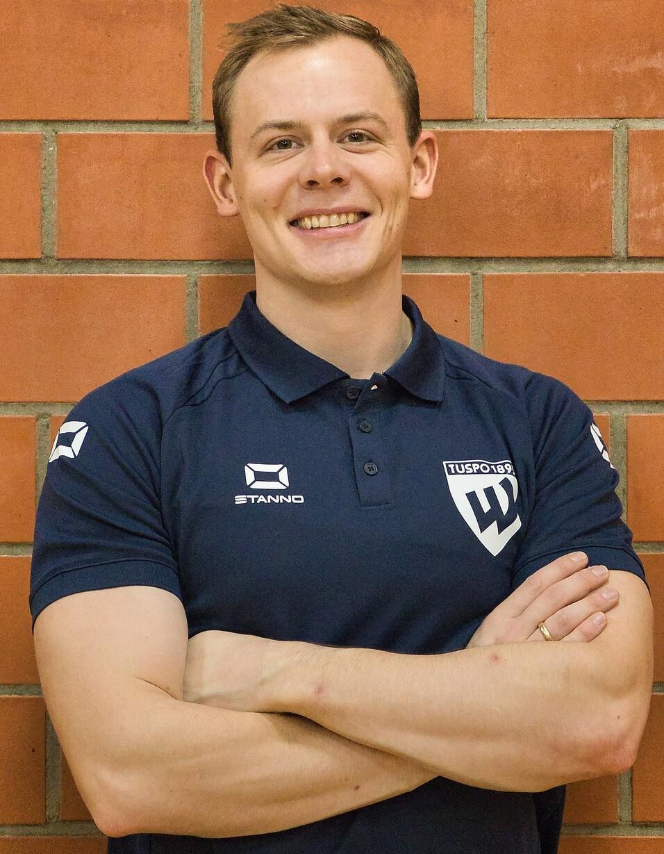 Mann mit kurzen Haaren und Smiling, trägt ein dunkelblaues Polo-Shirt, steht vor einer Backsteinwand.