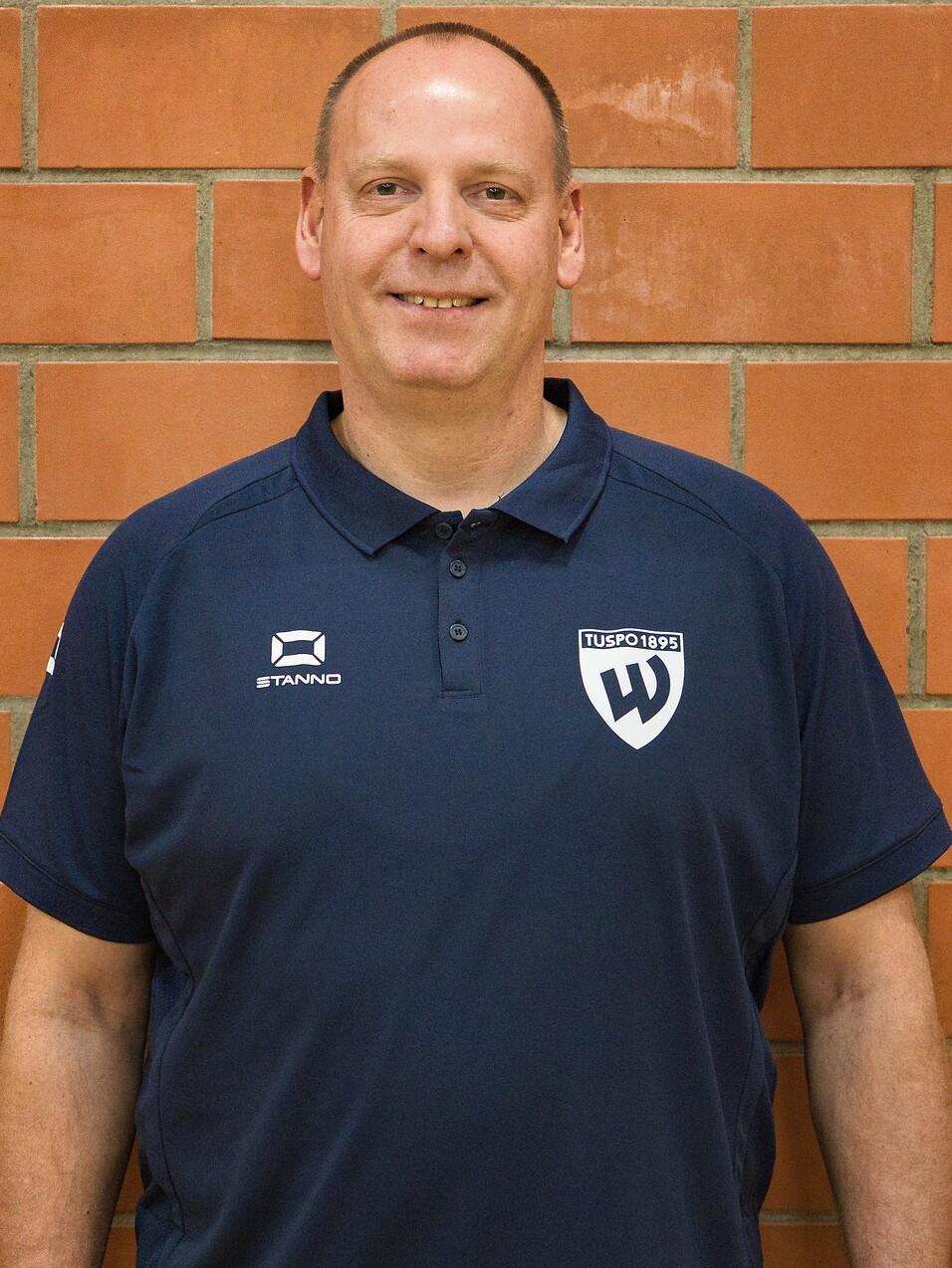 Mann mit kurzem Haar in sportlichem Polohemd vor einer roten Backsteinwand, lächelt freundlich in die Kamera.