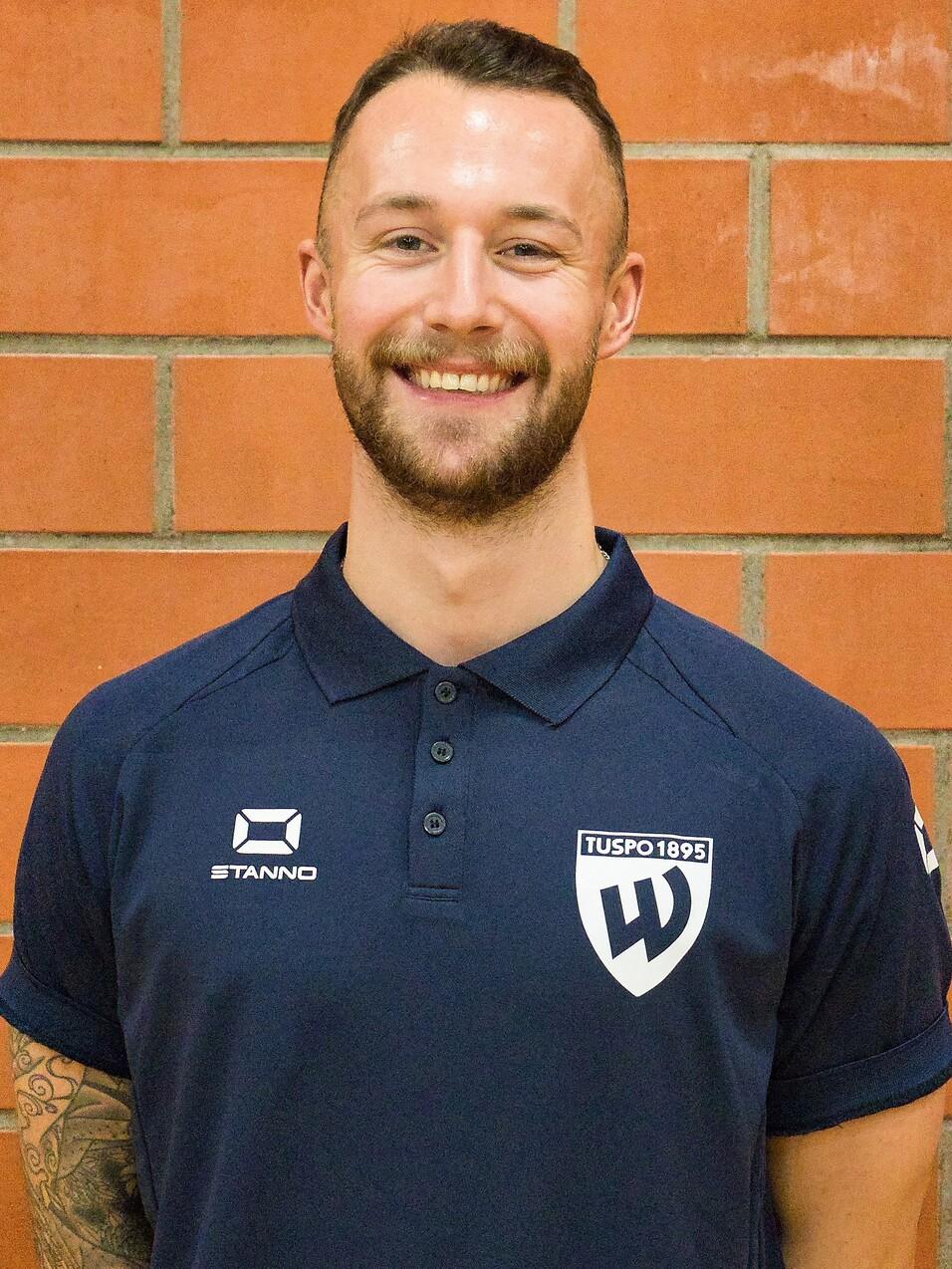 Porträt eines lächelnden Mannes mit Bart, in einem blauen Polo-Shirt mit Vereinslogo vor einer Backsteinwand.