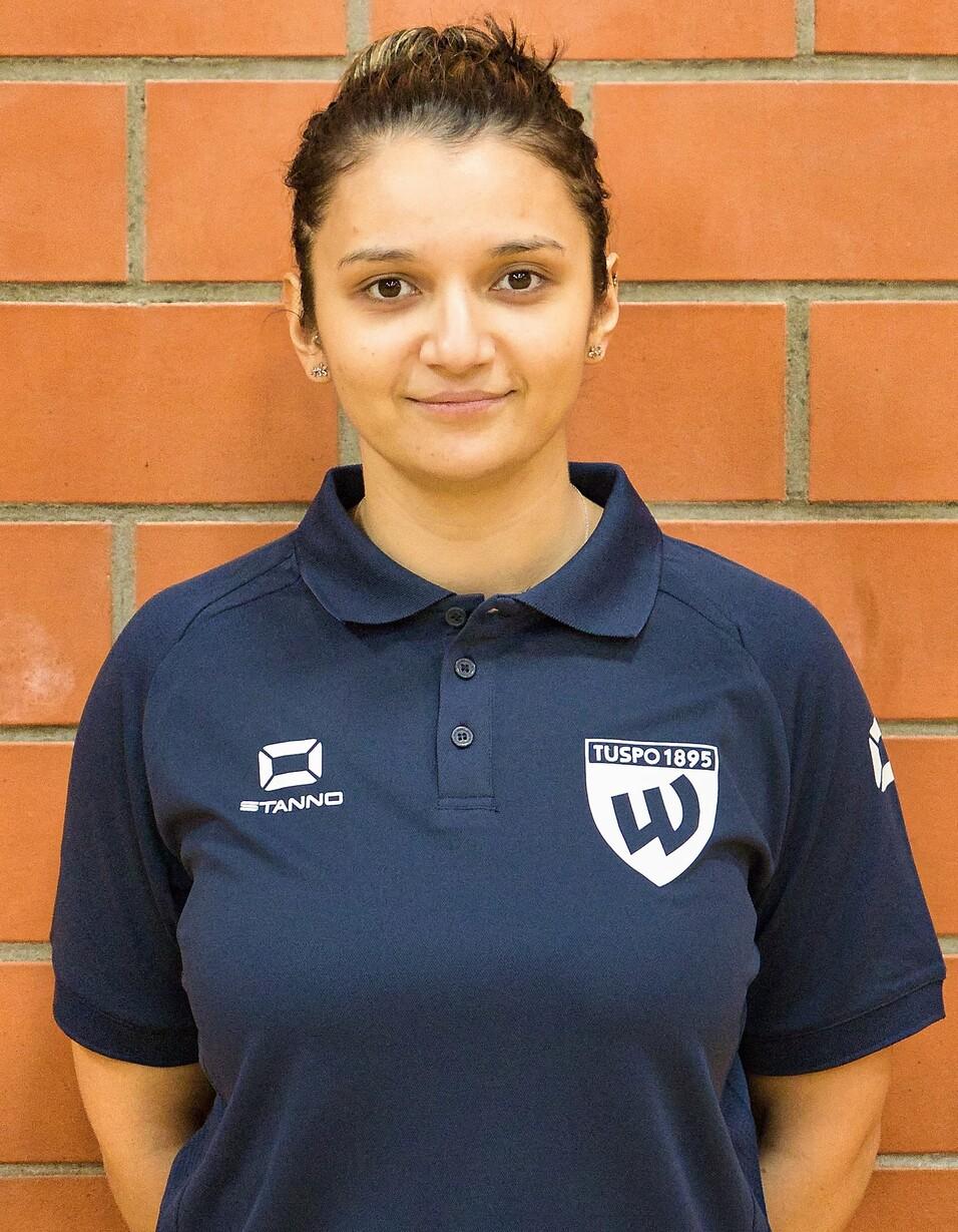 Porträt einer Person in einem dunkelblauen Poloshirt mit dem Logo von TUSPO 1895 vor einer Ziegellandwand.