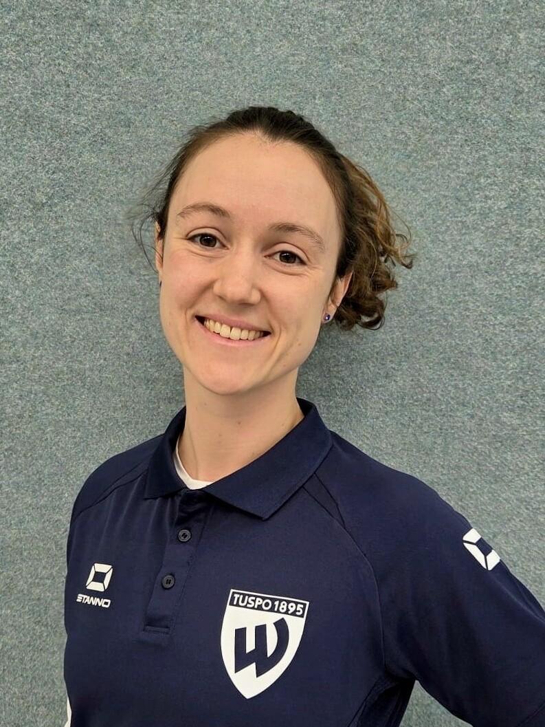 Porträt einer lächelnden Frau in einem dunkelblauen Polo mit dem Logo von TUSPO 1895 vor grauer Wand.