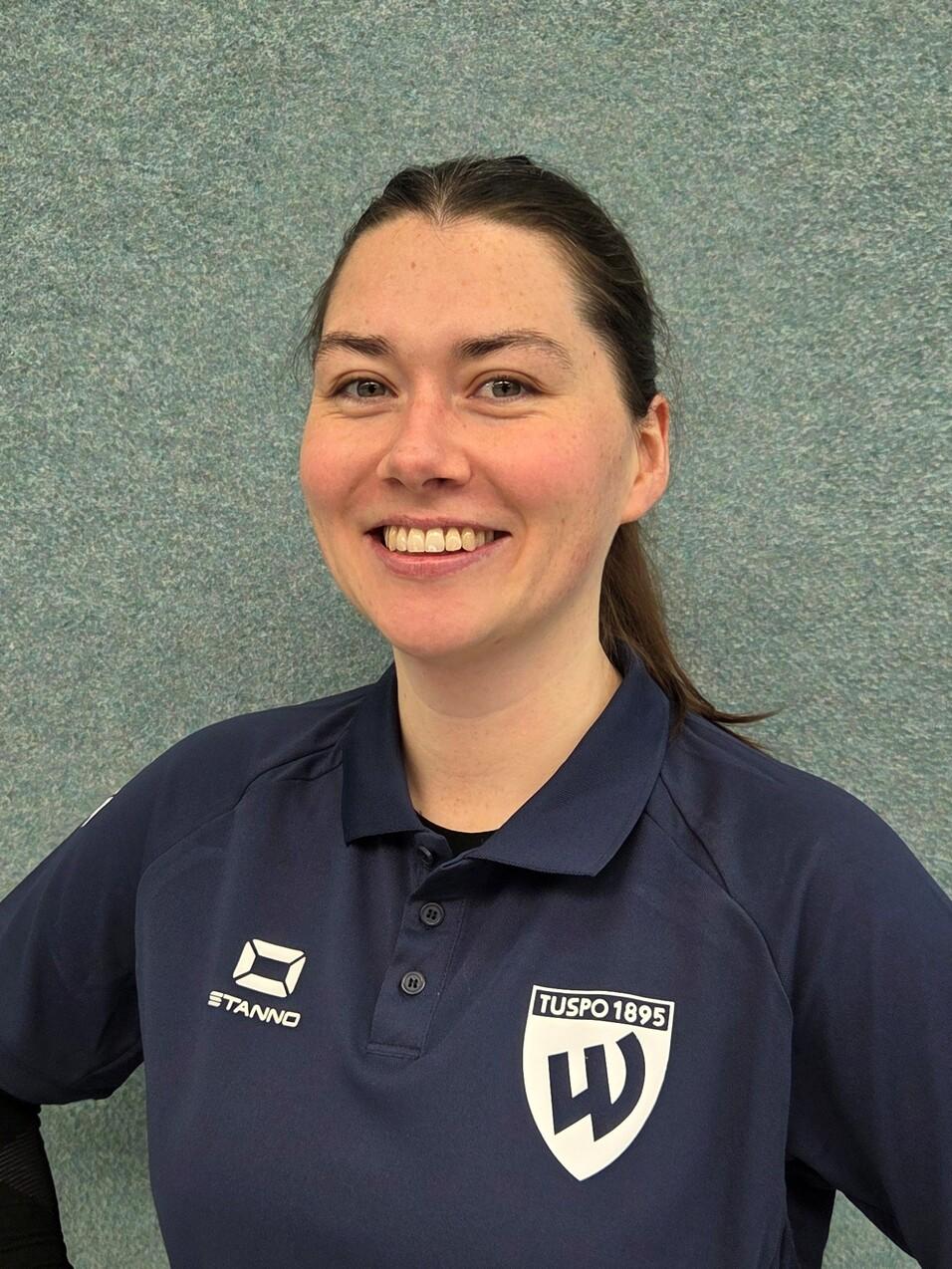 Lächelnde Frau in Sportkleidung mit Logo, steht vor grauer Wand mit den Händen in die Hüften gestützt.