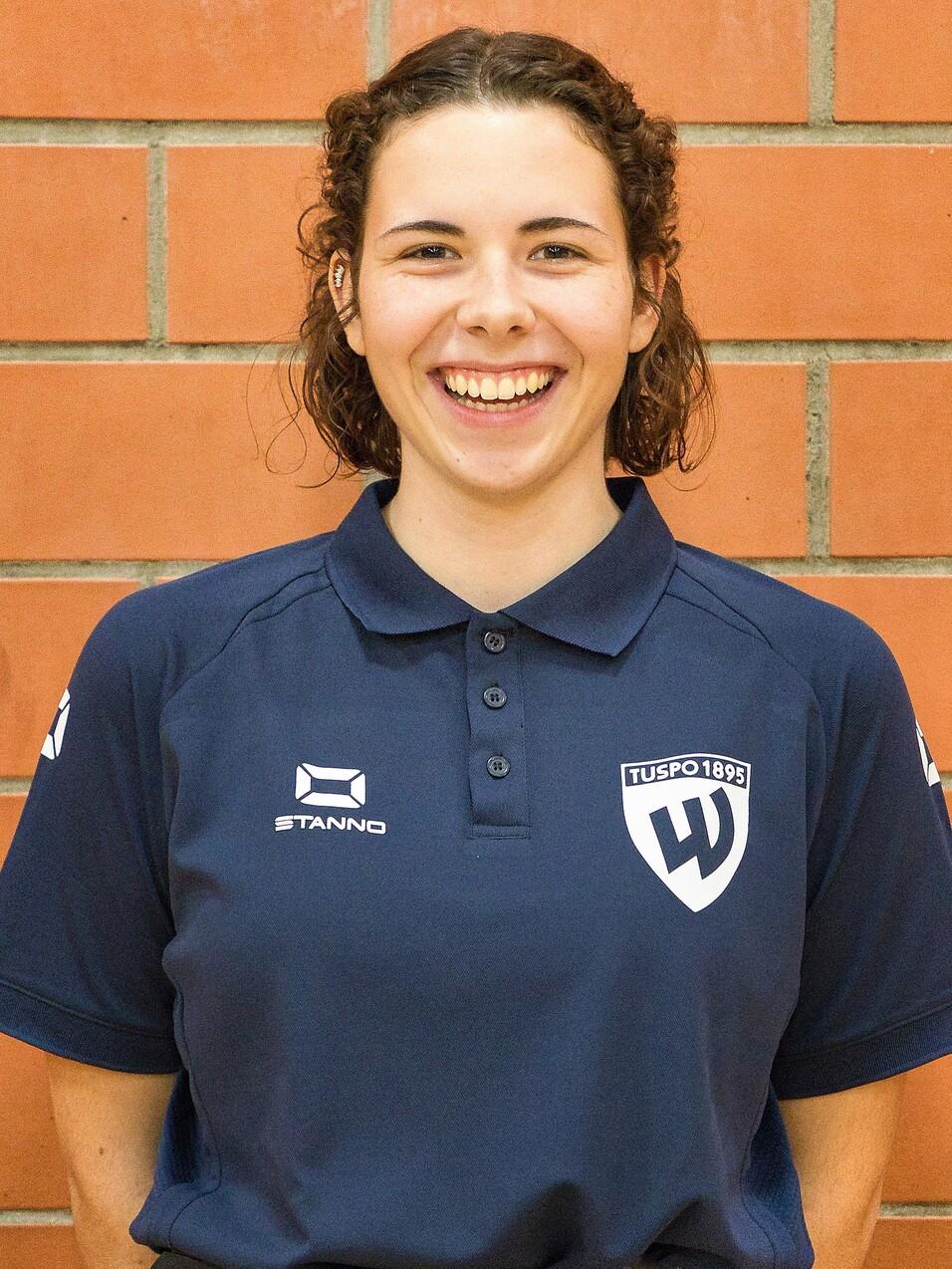 Lächelnde Frau mit lockigem Haar in dunkelblauem Poloshirt vor einer Ziegelwand, trägt ein Vereinslogo.