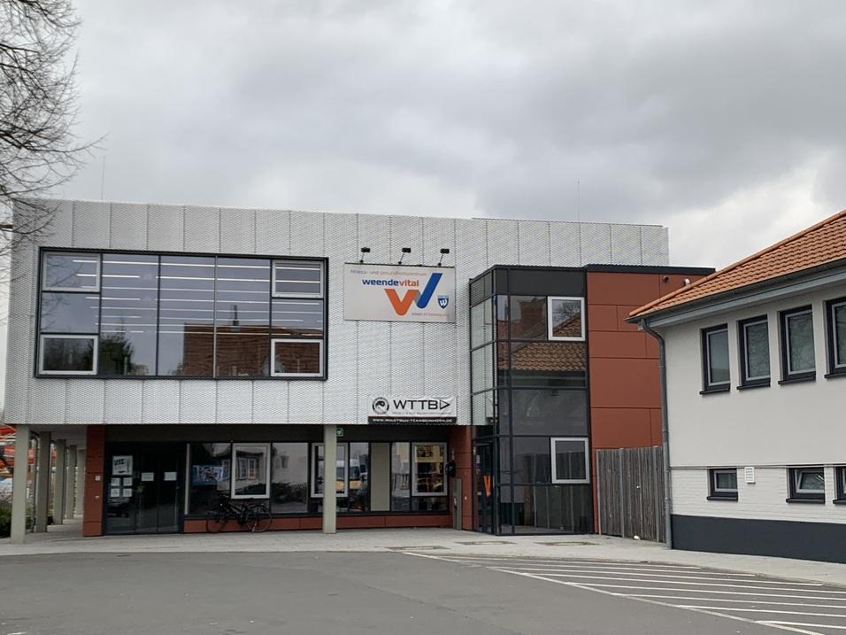Modernes Gebäude mit großem Fenster und dem Logo „vereinte-vital“ an der Fassade, angrenzendes Wohngebäude sichtbar.