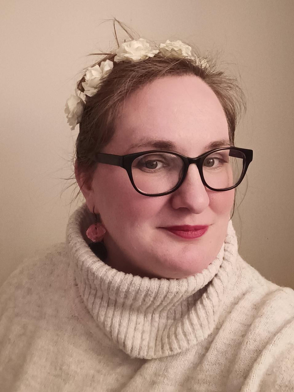 Frau mit Blumenkopfband, Brille und lächelndem Gesicht, trägt einen hohen, hellen Pullover. Hintergrund ist neutral.