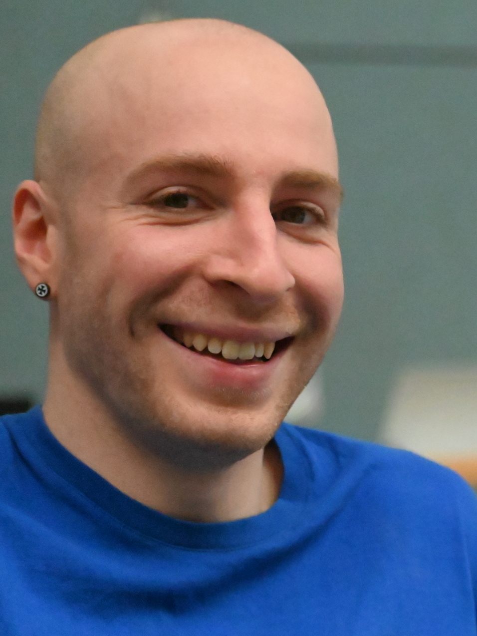 L&auml;chelnder Mann mit Glatze in blauer T-Shirt, sitzend vor grauem Hintergrund.