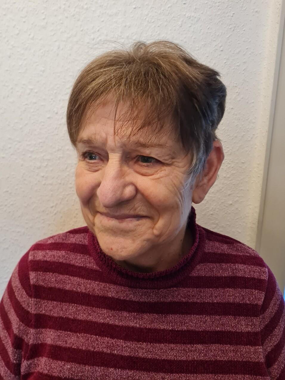 Porträt einer lächelnden älteren Frau mit kurzen Haaren, die einen gestreiften Pullover trägt, vor einer hellen Wand.