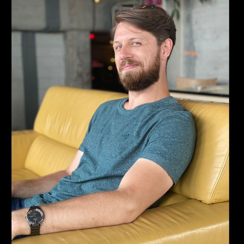 Mann mit Bart sitzt entspannt auf einer gelben Couch und lächelt in die Kamera.