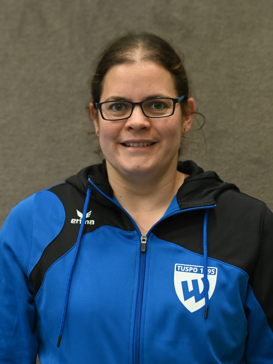 Frau mit Brille in blauer Sportjacke vor grauem Hintergrund, l&auml;chelt freundlich in die Kamera.