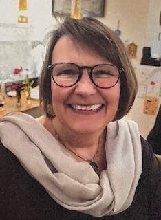 Lächelnde Frau mit Brille und Halstuch auf einem Hintergrund mit Dekoobjekten.