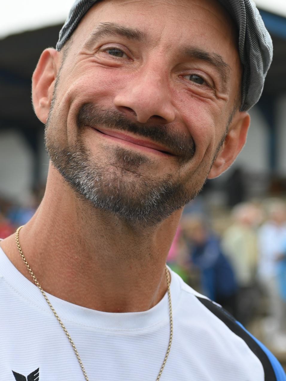 Portrait eines lächelnden Mannes in Sportkleidung mit grau-blauer Mütze, im Hintergrund verschwommene Zuschauer.