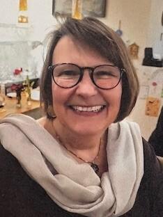 Lächelnde Frau mit Brille und Halstuch auf einem Hintergrund mit Dekoobjekten.