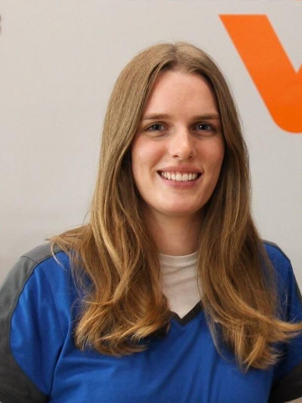 Lächelnde Frau mit langen, blonden Haaren, in blauer Arbeitskleidung, vor einem hellen Hintergrund mit Schriftzug.