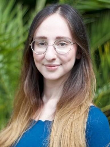 Porträt einer Frau mit langen, schulterlangen Haaren und Brille, vor einem Hintergrund aus grünen Pflanzen.