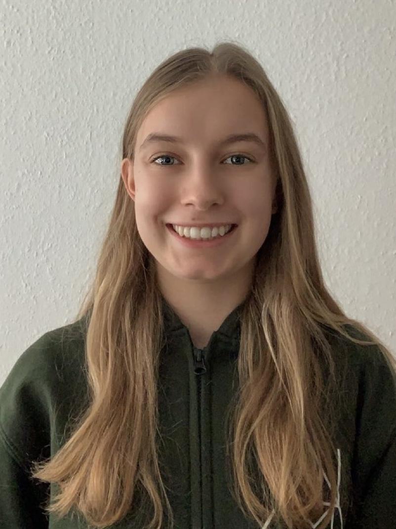 Lächelnde junge Frau mit langen, blonden Haaren in einem grünen Hoodie vor einer hellen Wand.