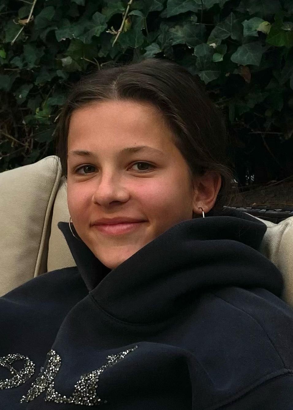 Porträt eines lächelnden Mädchens mit kurzen Haaren, das in einem schwarzen Kapuzenpullover auf einer Couch sitzt.