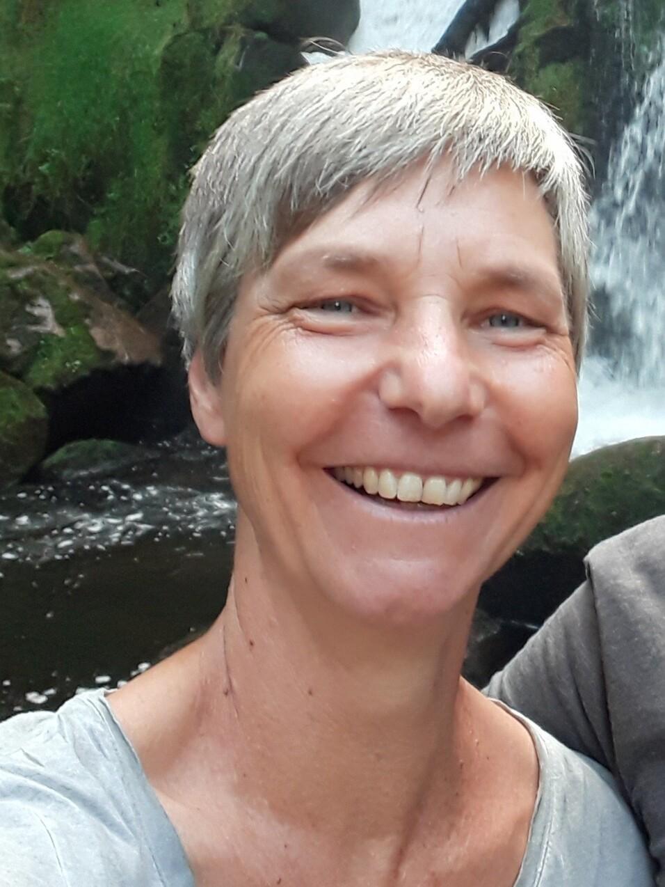 Lächelnde Frau mit kurzen grauen Haaren steht vor einem Wasserfall in einer grünen Umgebung.