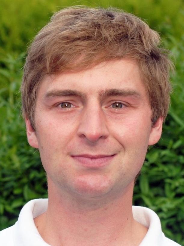 Porträt eines Mannes mit blondem Haar, der in einem weißen Poloshirt vor einer grünen Hecke steht.