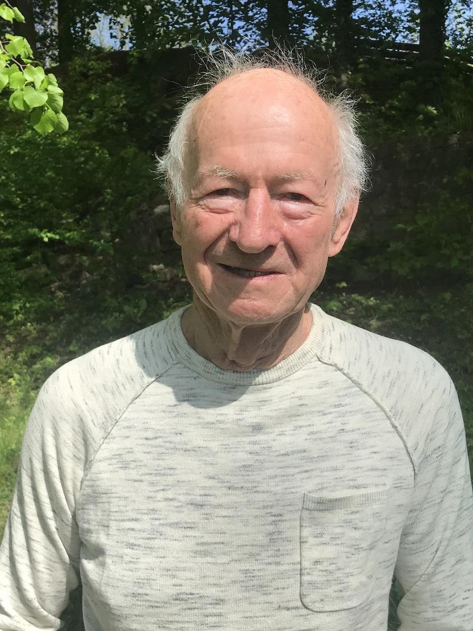 Älterer Mann steht vor grüner Hintergrundlandschaft, trägt einen hellen Pullover und lächelt freundlich in die Kamera.
