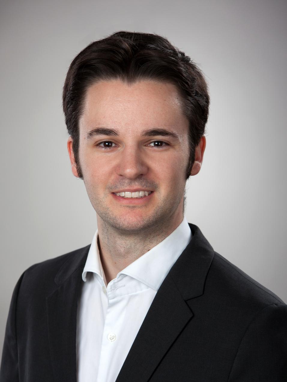 Porträt eines lächelnden Mannes in einem schwarzen Anzug mit weißem Hemd vor einem neutralen Hintergrund.