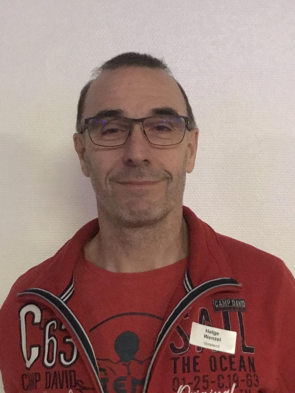 Mann mit Brille trägt roten Kapuzenpullover und lächelt vor einem hellen Hintergrund. Namensschild am Oberteil sichtbar.