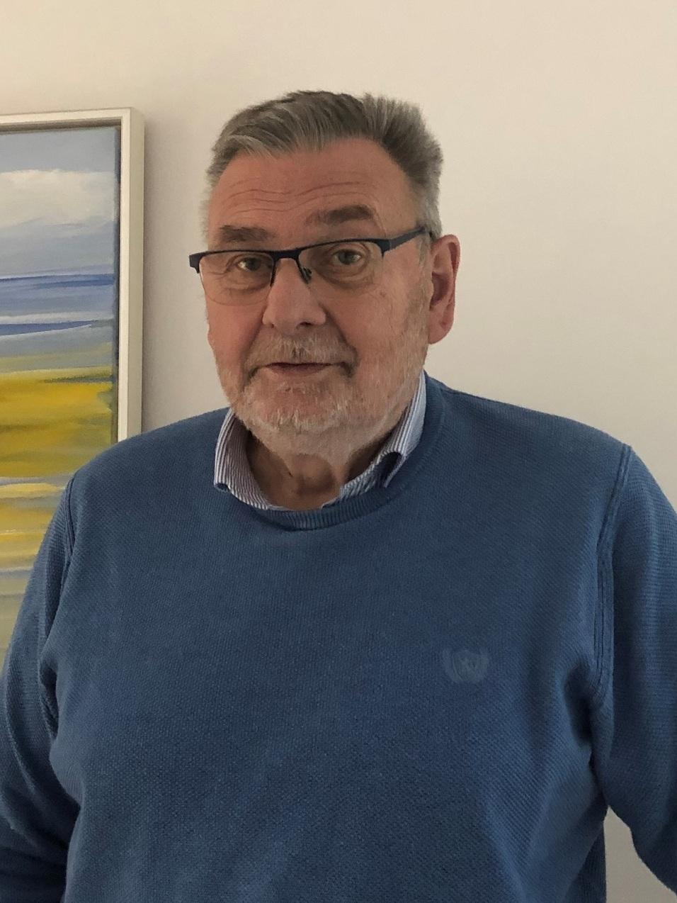 Älterer Mann mit Brille in blauer Pulloverjacke steht neben einem abstrakten Gemälde an einer Wand.