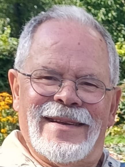 &Auml;lterer Mann mit Brille und grauem Bart, l&auml;chelt vor einer gr&uuml;nen Umgebung mit bunten Blumen.