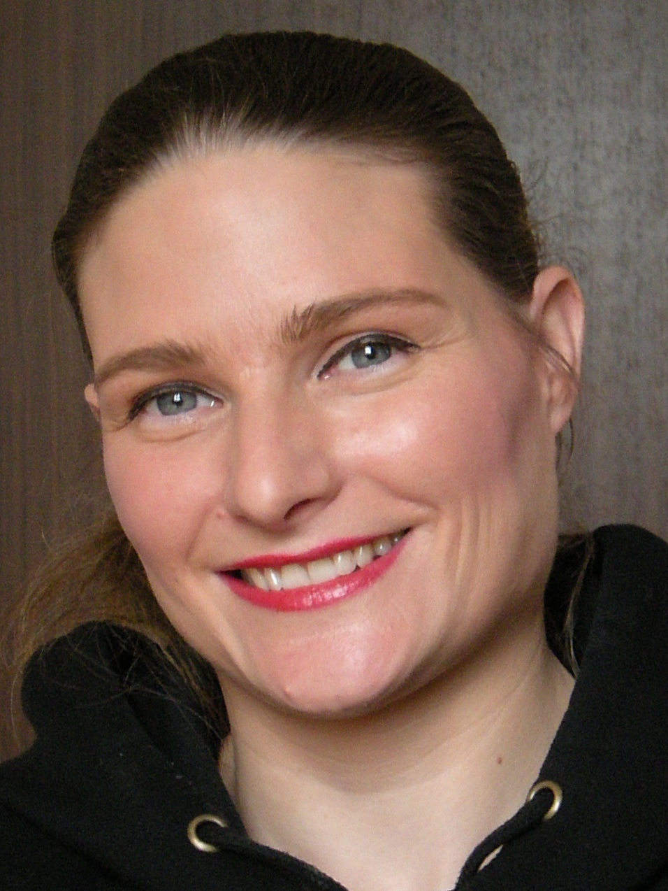 L&auml;chelnde Frau mit langen, braunen Haaren und rotem Lippenstift, tr&auml;gt einen schwarzen Kapuzenpullover vor einem Holz-Hintergrund.