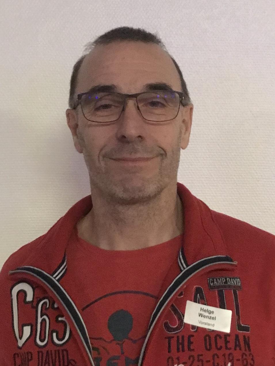 Mann mit Brille trägt roten Kapuzenpullover und lächelt vor einem hellen Hintergrund. Namensschild am Oberteil sichtbar.