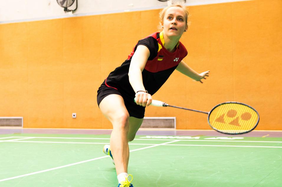 Spielerin schlägt einen Badmintonball auf einer grünen Spielfläche in einer Sporthalle mit orangefarbenen Wänden.