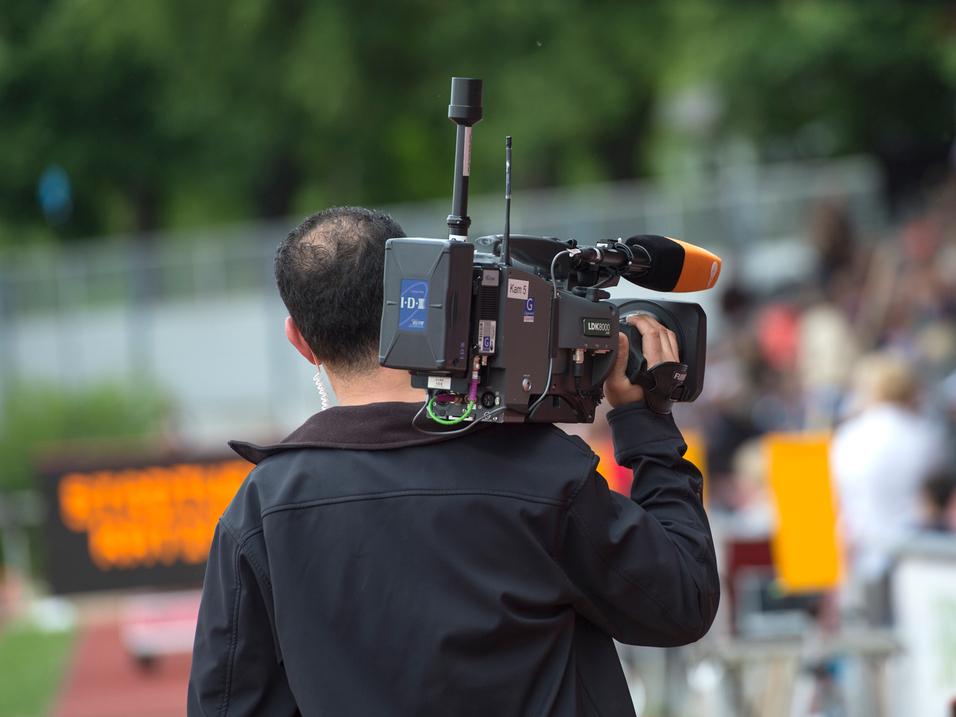 Kameramann filmt bei einer Sportveranstaltung, Hintergrund zeigt eine Zuschauertribüne und eine Zeitmessanzeige.
