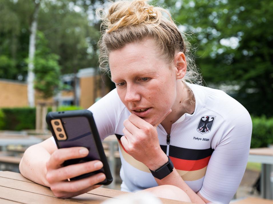 Athletin im weißen Trikot mit schwarzem und rotem Streifen schaut konzentriert auf ihr Smartphone in einer Außenanlage.